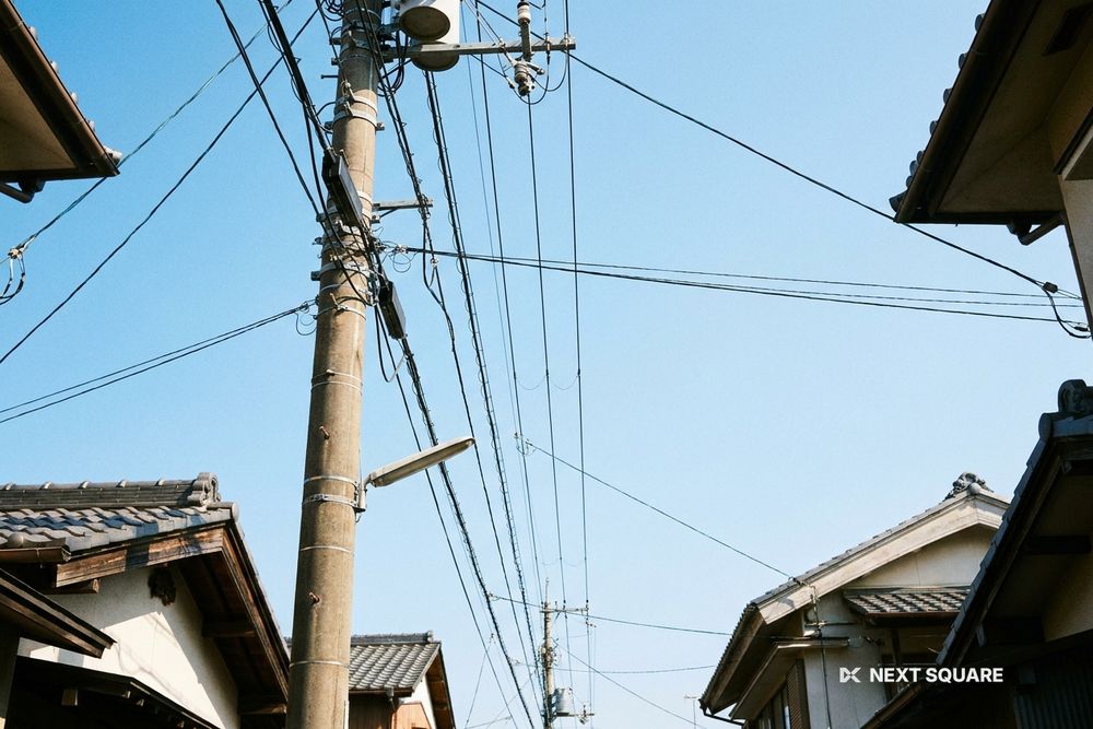 電柱から家々へ張られた架線を見上げた景色、青空と瓦屋根(文字や人物は写っていない)